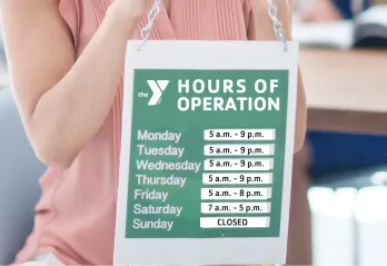 Woman holding up a sign with  hours of operation for YMCA branch hours