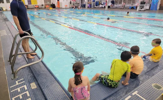 Man standing on the edge of the 4 feet end of pool with children sitting on the edge of pool