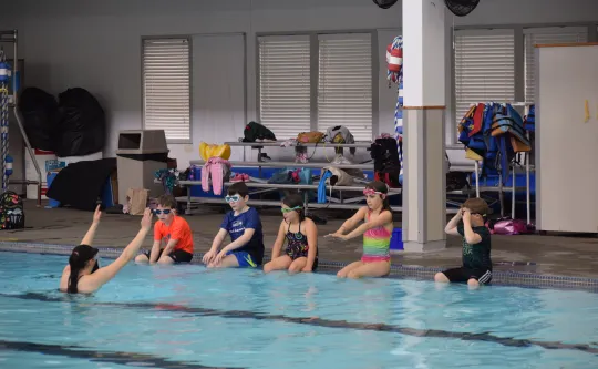 Instructor in the pool with children sitting on the side of the pool learning how to dive into pool