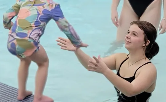 A teen in the shallow poo reaching her hands out to a child who is going to jump into the pool